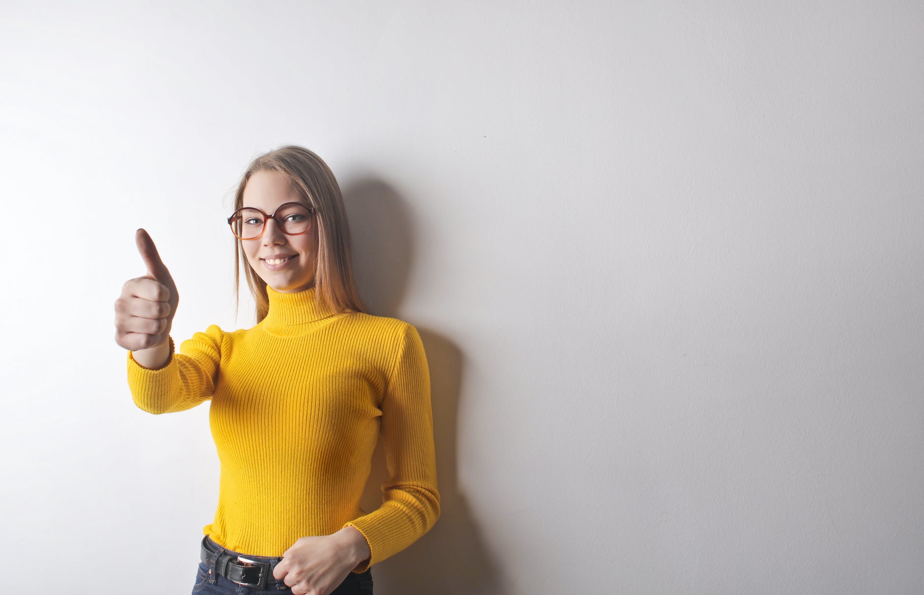 photo of woman in yellow turtleneck sweater blue denim jeans and glasses giving the thumbs up